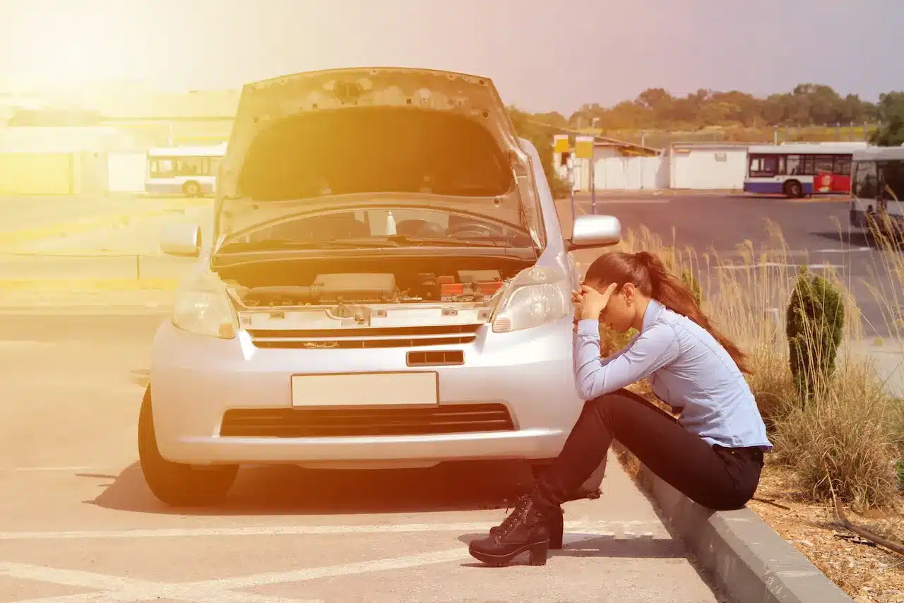 A woman sits on the ground beside a broken-down car