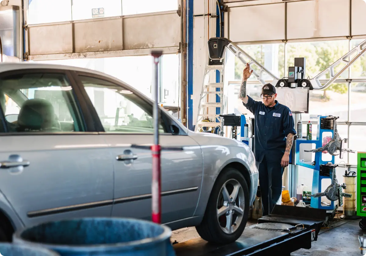 A mechanic is standing in front of a car in a garage, with shelves and tools in the background