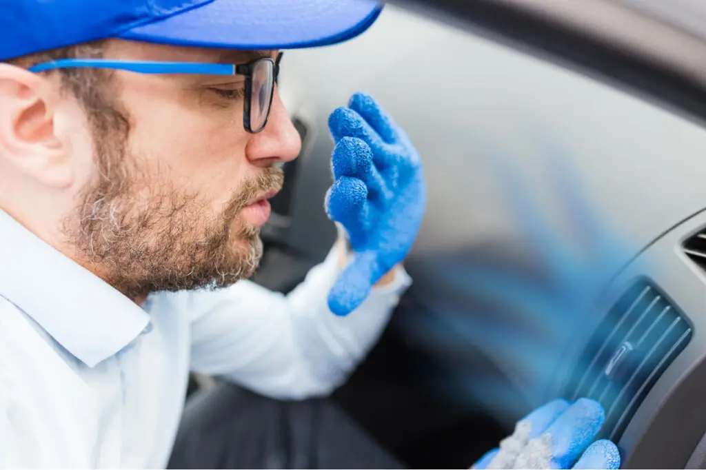 A man wearing gloves and a blue cap is focused on checking if there is air blowing from the car's AC unit