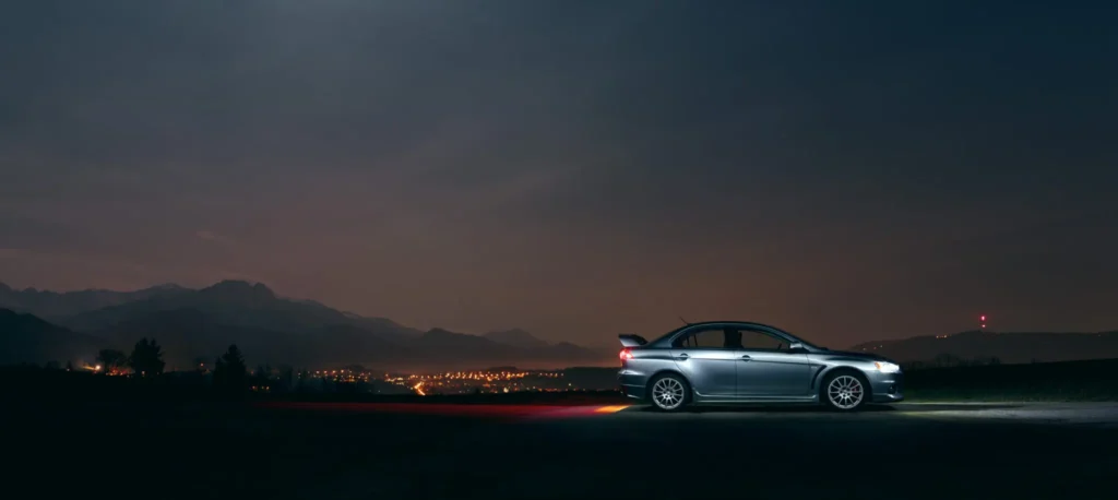 A car parked on a dimly lit road at night, surrounded by shadows and illuminated by distant streetlights.